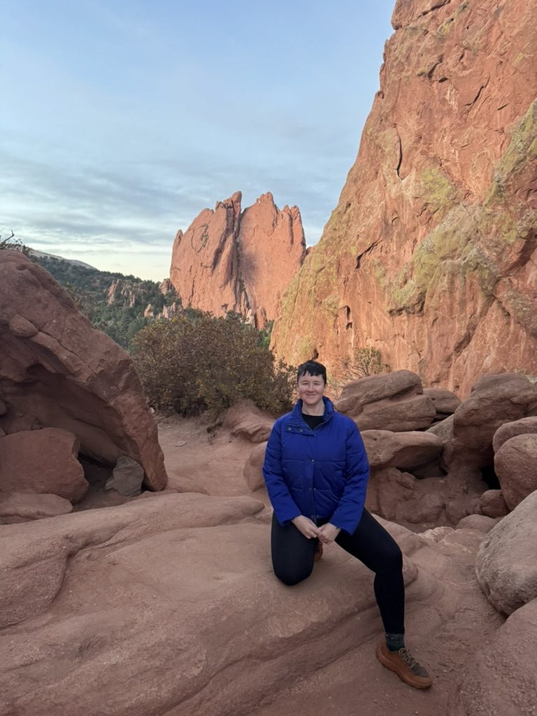 Virginia in a blue puffer jacket, kneeling on red rocks in Colorado at sunrise.
